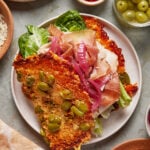 A plate with a sandwich featuring a golden, crispy cheese and green olive crust, filled with lettuce, pickled red onions, prosciutto, and creamy cheese. Small bowls of green olives and red sauce are nearby.