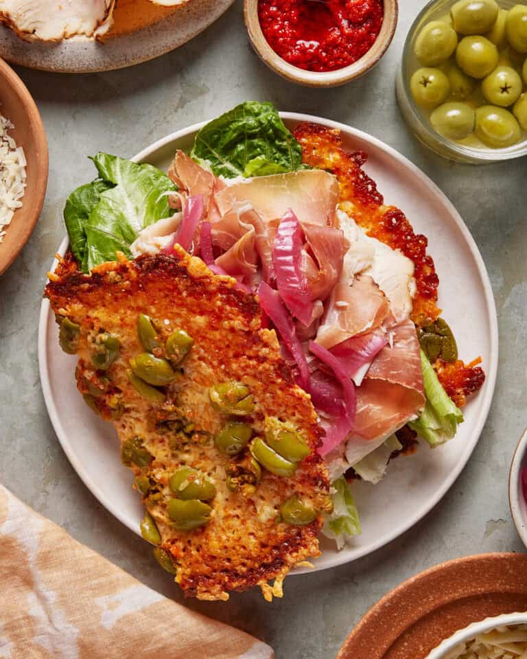 A plate with a sandwich featuring a golden, crispy cheese and green olive crust, filled with lettuce, pickled red onions, prosciutto, and creamy cheese. Small bowls of green olives and red sauce are nearby.