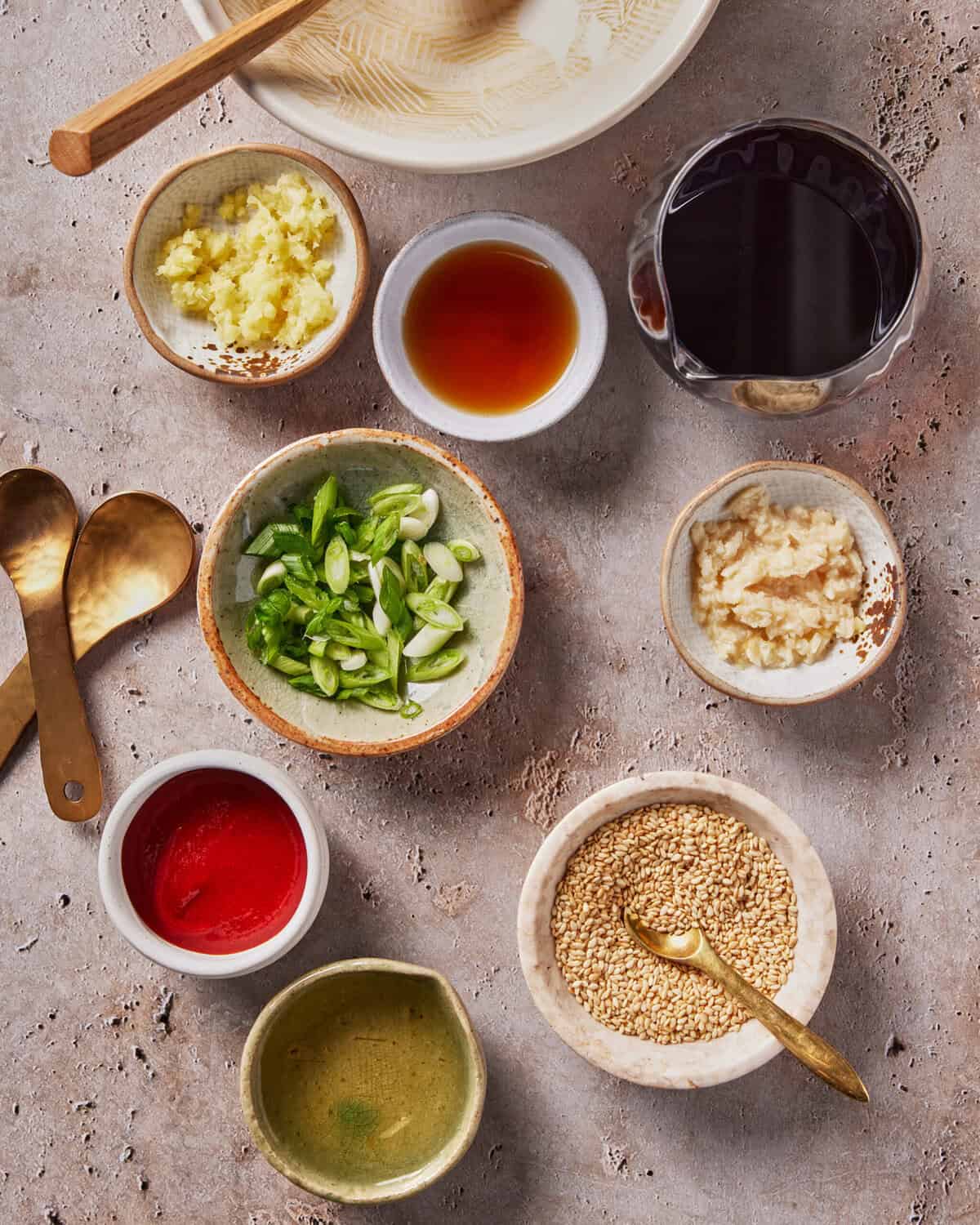 A flat lay of various small bowls holding chopped green onions, minced garlic, sesame seeds, grated ginger, red chili paste, soy sauce, and a golden liquid, arranged on a stone surface with two brass spoons.