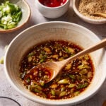 A bowl of soy-based dipping sauce with sliced green onions and sesame seeds, stirred with a wooden spoon. Surrounding the bowl are small dishes of sesame seeds, sliced green onions, and red sauce.