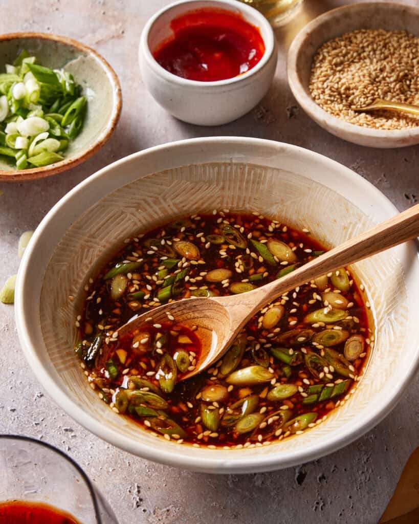 A bowl of soy-based dipping sauce with sliced green onions and sesame seeds, stirred with a wooden spoon. Surrounding the bowl are small dishes of sesame seeds, sliced green onions, and red sauce.