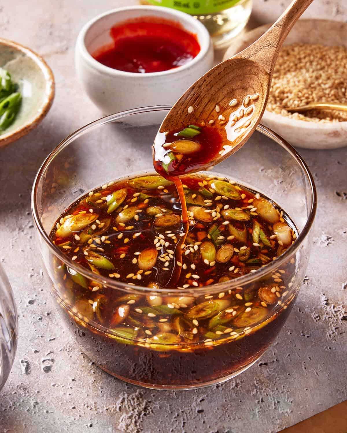 A glass bowl filled with a dark, glossy sauce containing chopped green onions and sesame seeds, with a wooden spoon lifting some sauce. In the background are small bowls of red sauce and sesame seeds.