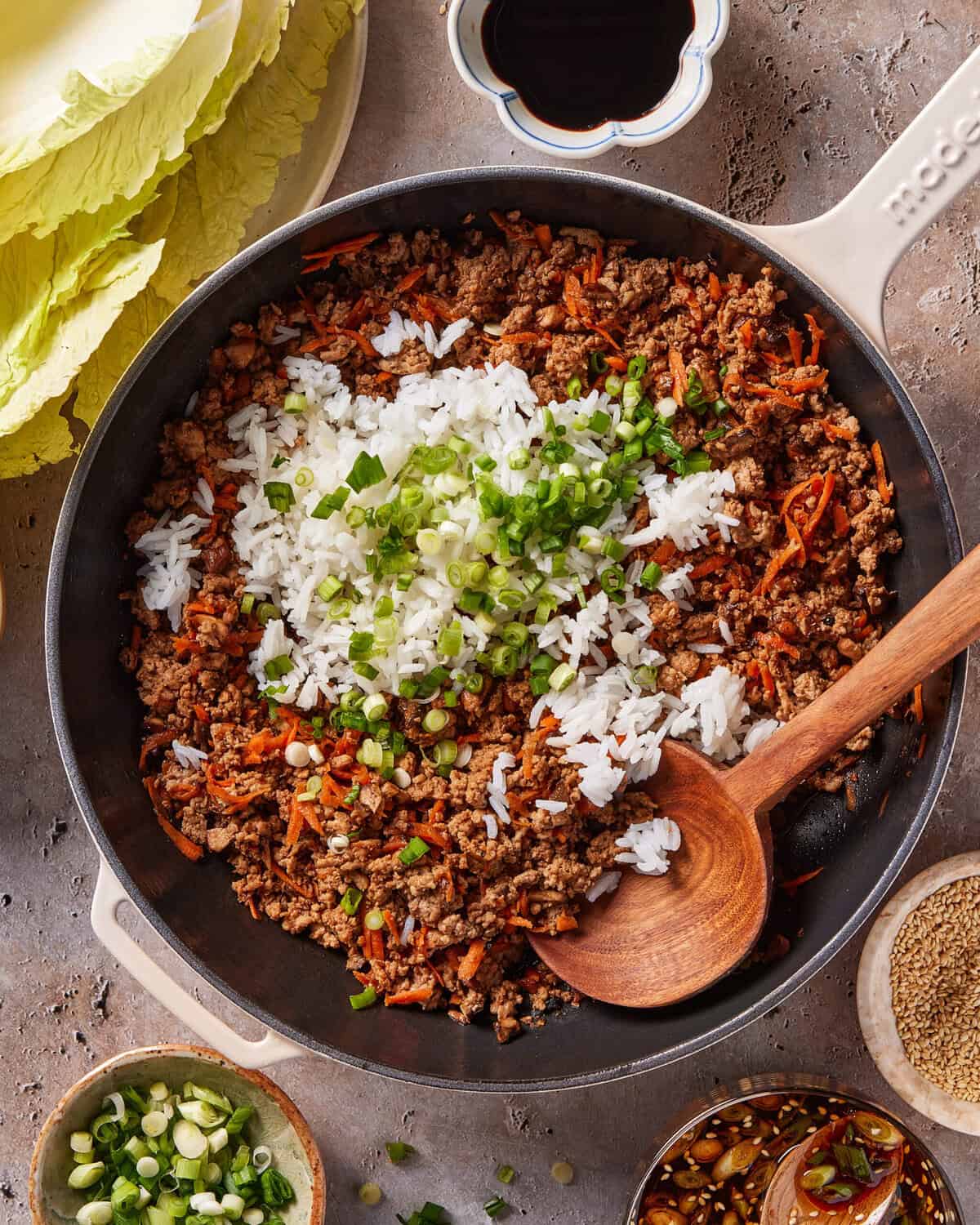 A skillet filled with cooked ground meat, shredded carrots, white rice, and chopped green onions, with a wooden spoon resting inside. Surrounding the skillet are cabbage leaves, sauce, and small bowls of seasonings.