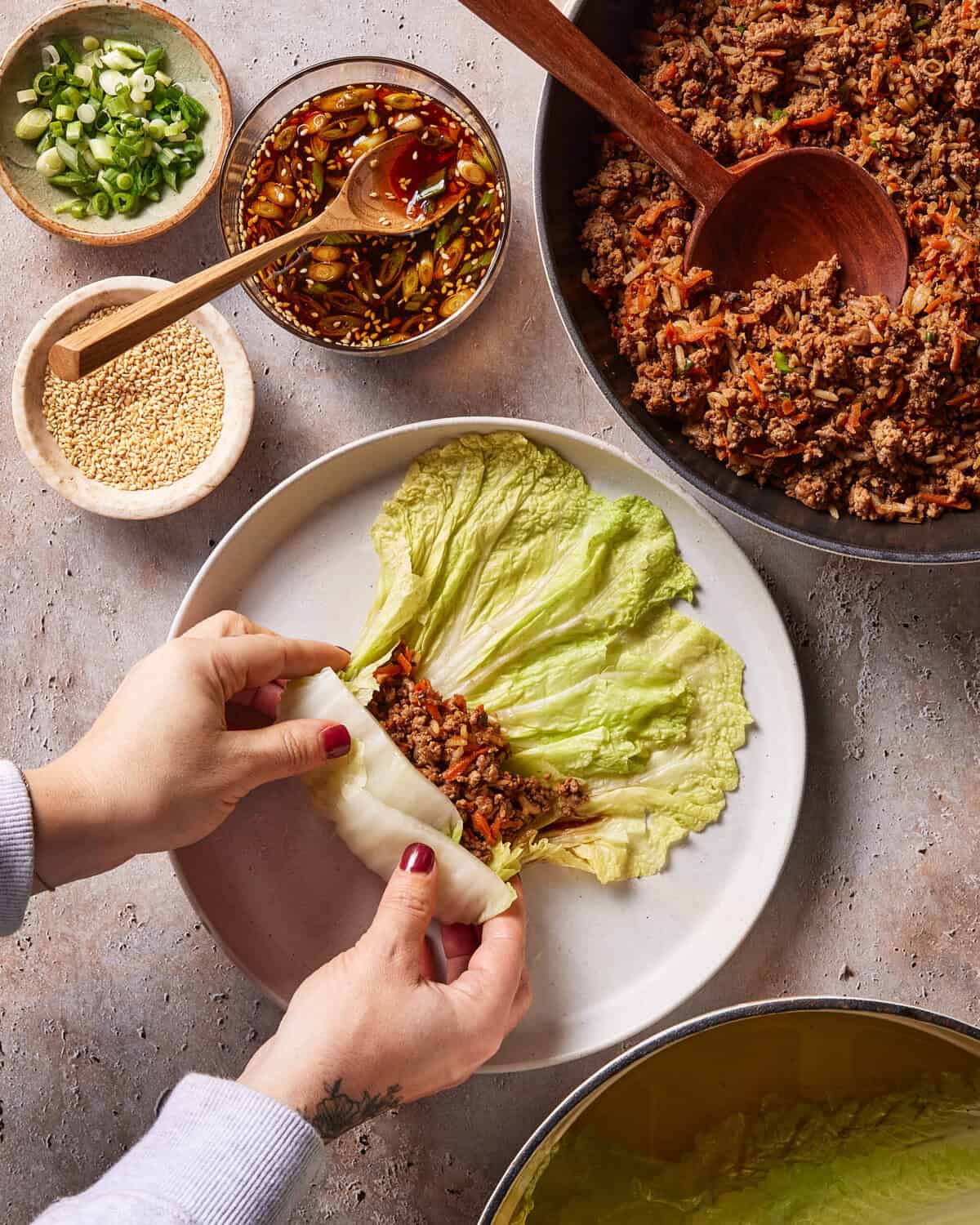 A person wraps cooked ground meat and vegetables in a large lettuce leaf on a plate. Nearby are bowls of sauce, sesame seeds, chopped green onions, and a pan of the meat mixture.