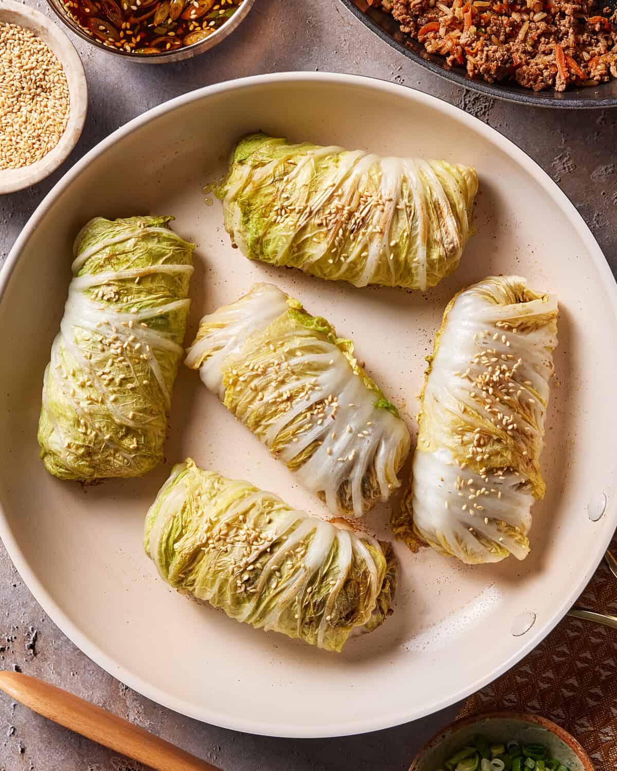 Five cabbage rolls filled with a savory mixture are arranged in a light-colored pan, topped with sesame seeds. Surrounding the pan are small bowls containing sesame seeds, a dark dipping sauce, and minced meat.