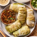 A plate of cabbage rolls sprinkled with sesame seeds, served with a bowl of soy-based dipping sauce garnished with chopped green onions and sesame seeds. Small bowls of sesame seeds and green onions are nearby.