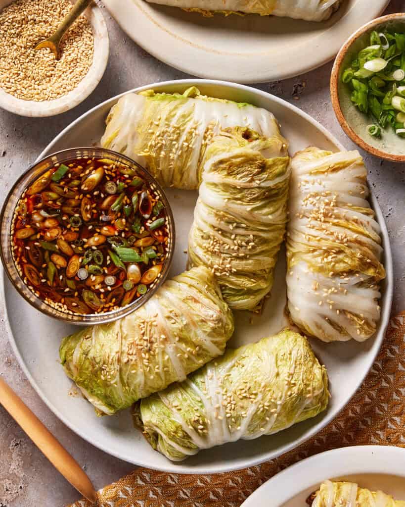 A plate of cabbage rolls sprinkled with sesame seeds, served with a bowl of soy-based dipping sauce garnished with chopped green onions and sesame seeds. Small bowls of sesame seeds and green onions are nearby.