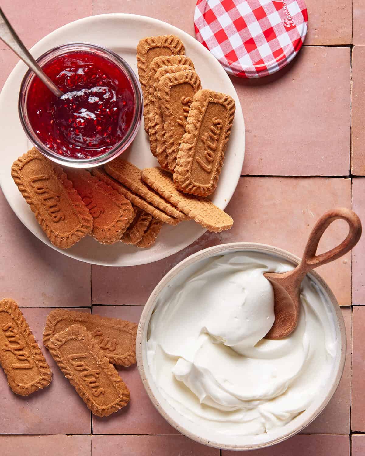 A bowl of creamy white yogurt with a wooden spoon, a plate of Lotus Biscoff cookies, and a glass jar of red fruit jam with a spoon, all arranged on a tiled surface.