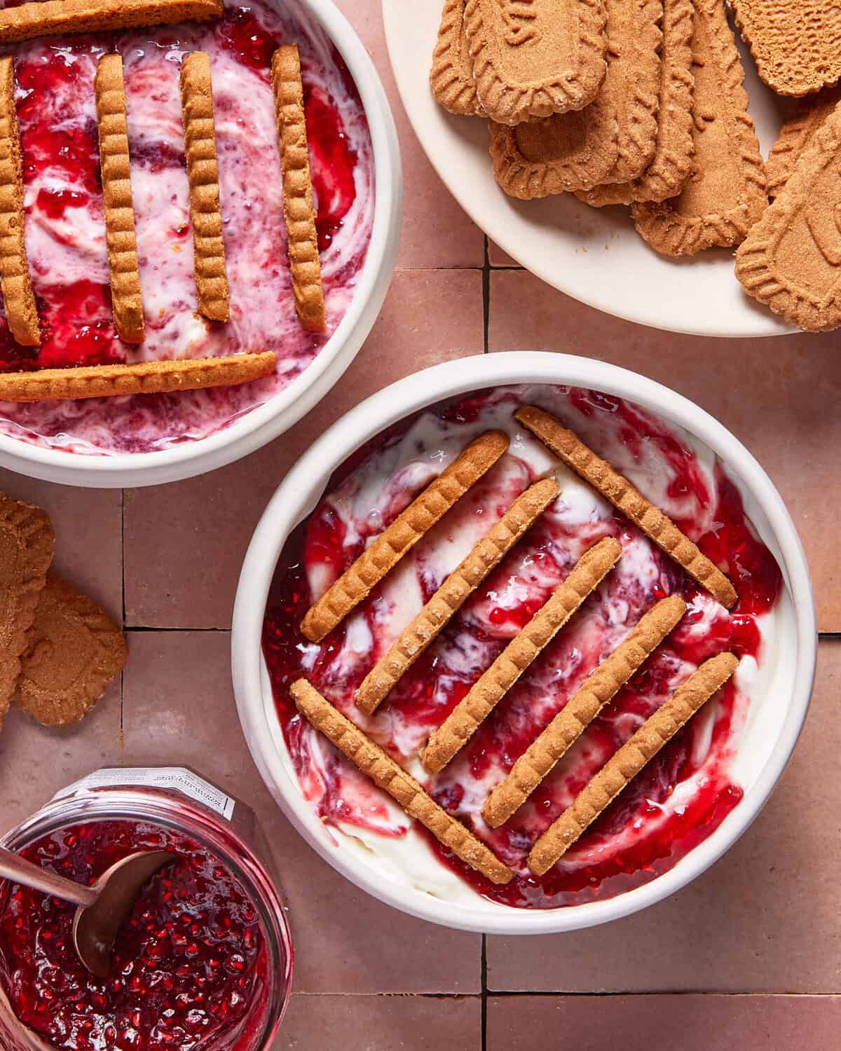 Two bowls of creamy yogurt swirled with raspberry jam, topped with rows of rectangular cookies. Nearby are a plate of cookies and a jar of raspberry jam on a tiled surface.