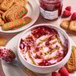 A bowl of yogurt topped with raspberry jam and cookie crumbs on a pink surface, surrounded by fresh raspberries, a plate of Biscoff cookies, a spoon with jam, and an open jar of raspberry preserves.