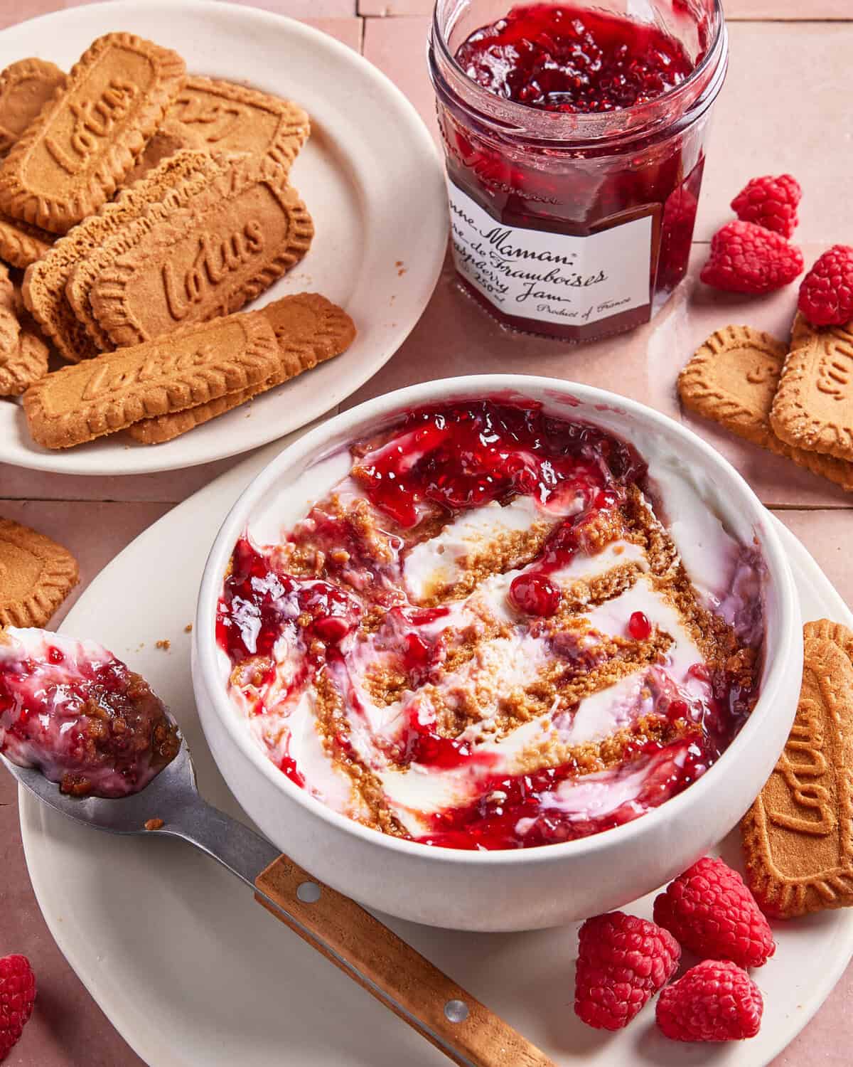 A bowl of yogurt topped with raspberry jam and cookie crumbs on a pink surface, surrounded by fresh raspberries, a plate of Biscoff cookies, a spoon with jam, and an open jar of raspberry preserves.