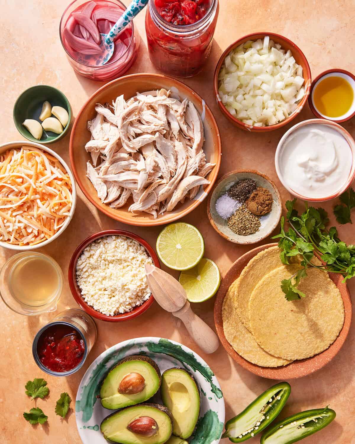 A flat lay of taco ingredients including shredded chicken, diced onions, avocado halves, shredded cheese, sliced jalapeño, corn tortillas, lime halves, chopped cilantro, sour cream, pickled onions, garlic cloves, and assorted spices.