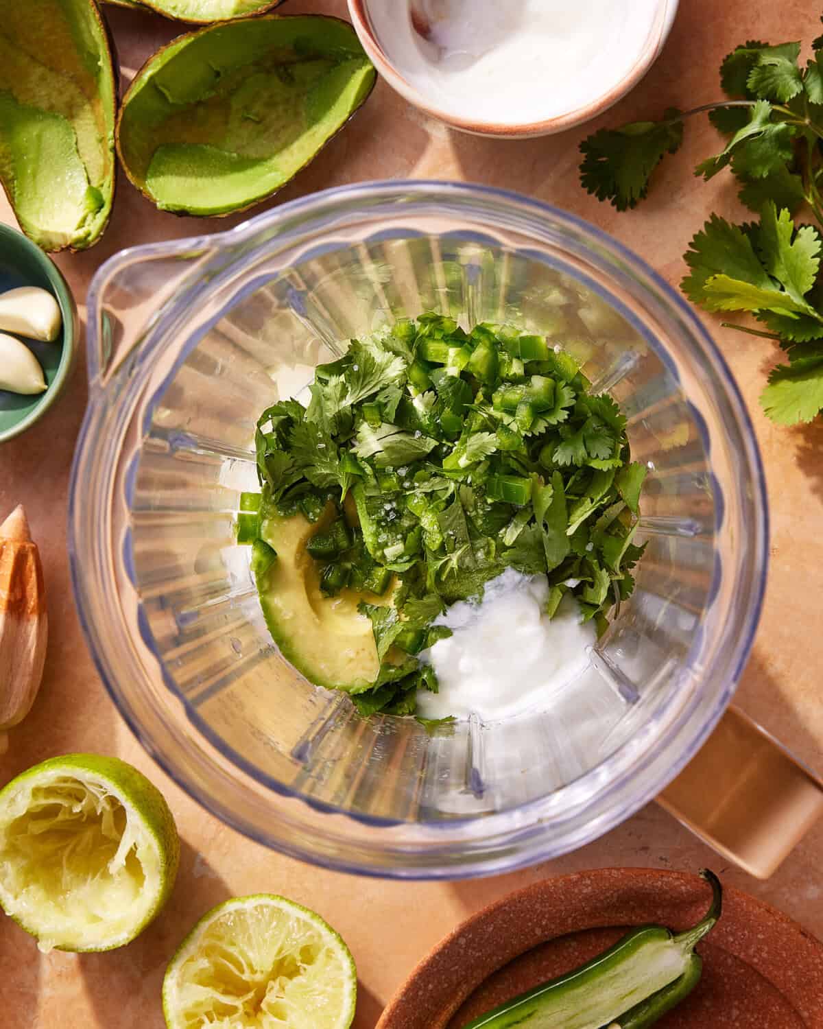 A blender filled with avocado, sliced jalapeño, cilantro, and yogurt sits on a countertop surrounded by lime halves, avocado skins, garlic, and fresh cilantro.