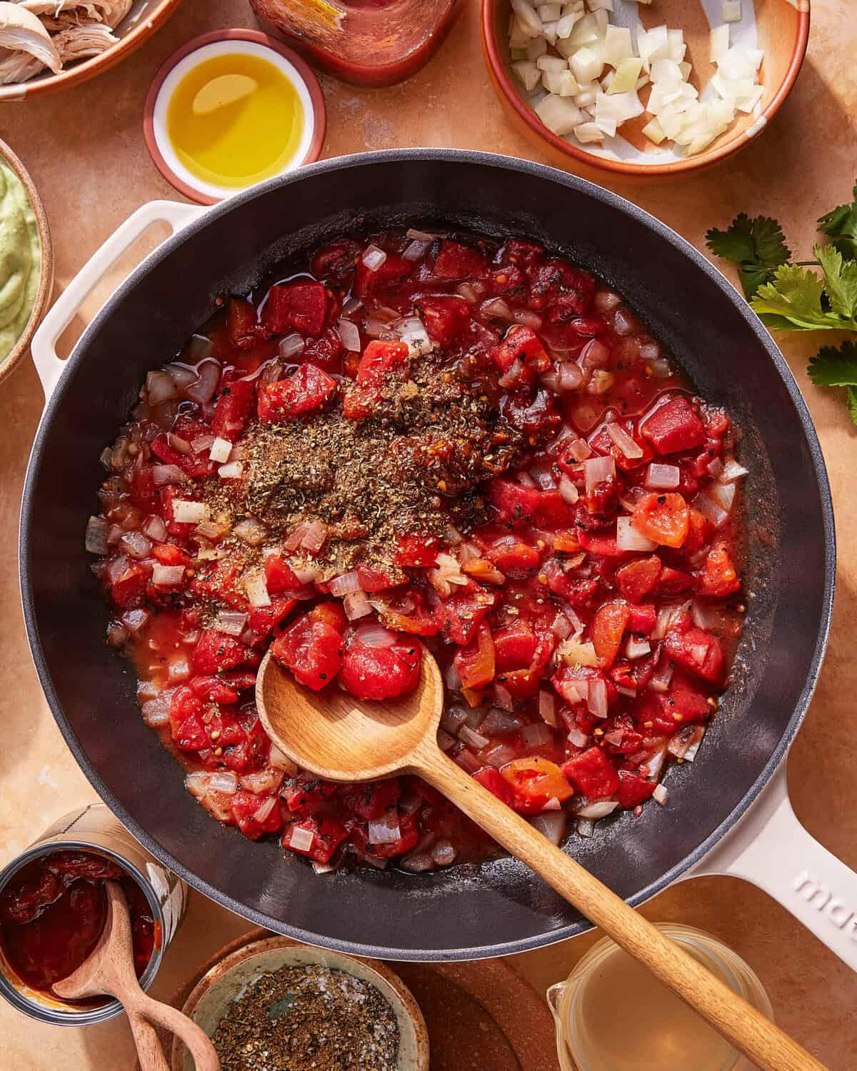 A skillet filled with chopped tomatoes, onions, and herbs being stirred with a wooden spoon. Surrounding the skillet are bowls containing olive oil, chopped onions, spices, and fresh cilantro.