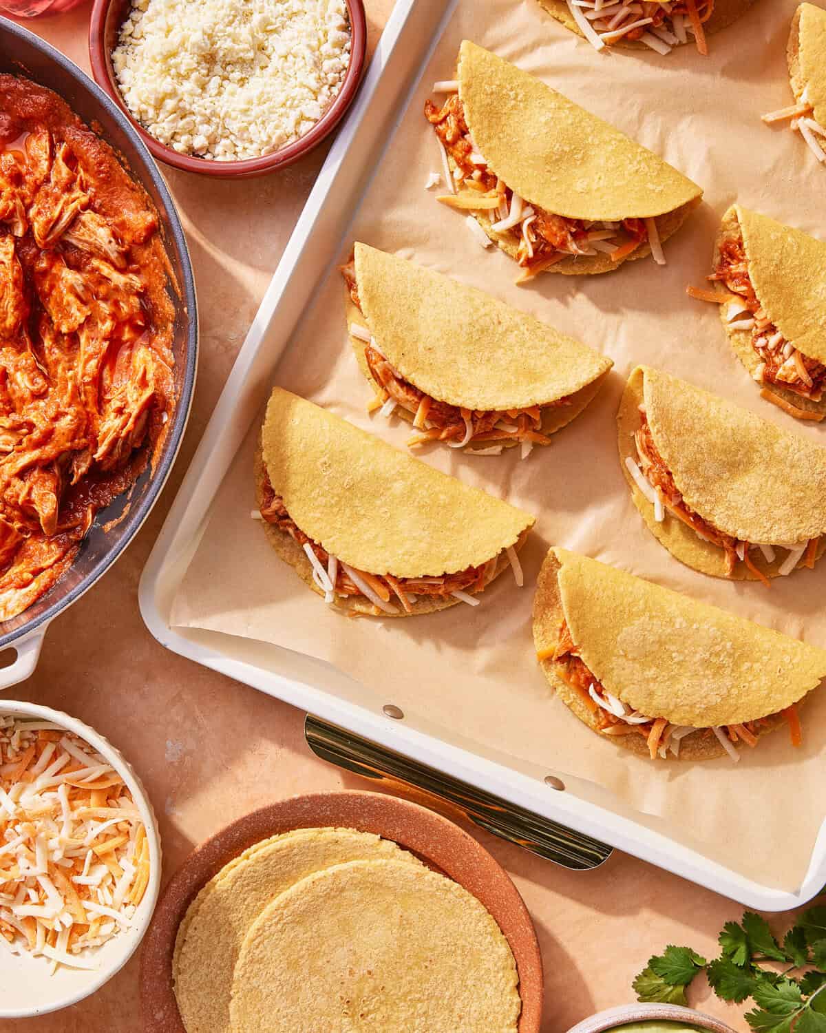 Corn tortillas filled with shredded chicken and cheese are arranged on a parchment-lined baking tray. Nearby are bowls of shredded chicken, cheese, crumbly cheese, extra tortillas, and fresh cilantro.