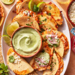 A plate of crispy tacos filled with cheese and garnished with cilantro and cheese, served with lime wedges and a bowl of creamy green dipping sauce in the center. A drink and small bowl of cheese are nearby.