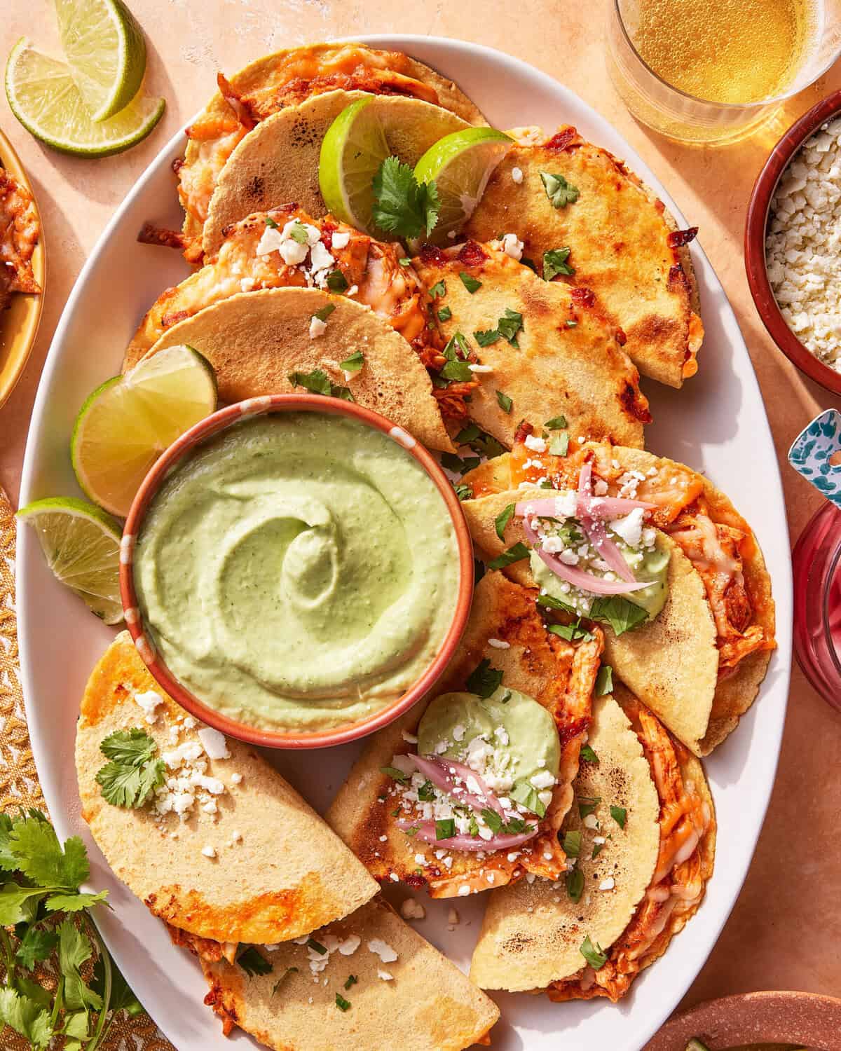 A plate of crispy tacos filled with cheese and garnished with cilantro and cheese, served with lime wedges and a bowl of creamy green dipping sauce in the center. A drink and small bowl of cheese are nearby.
