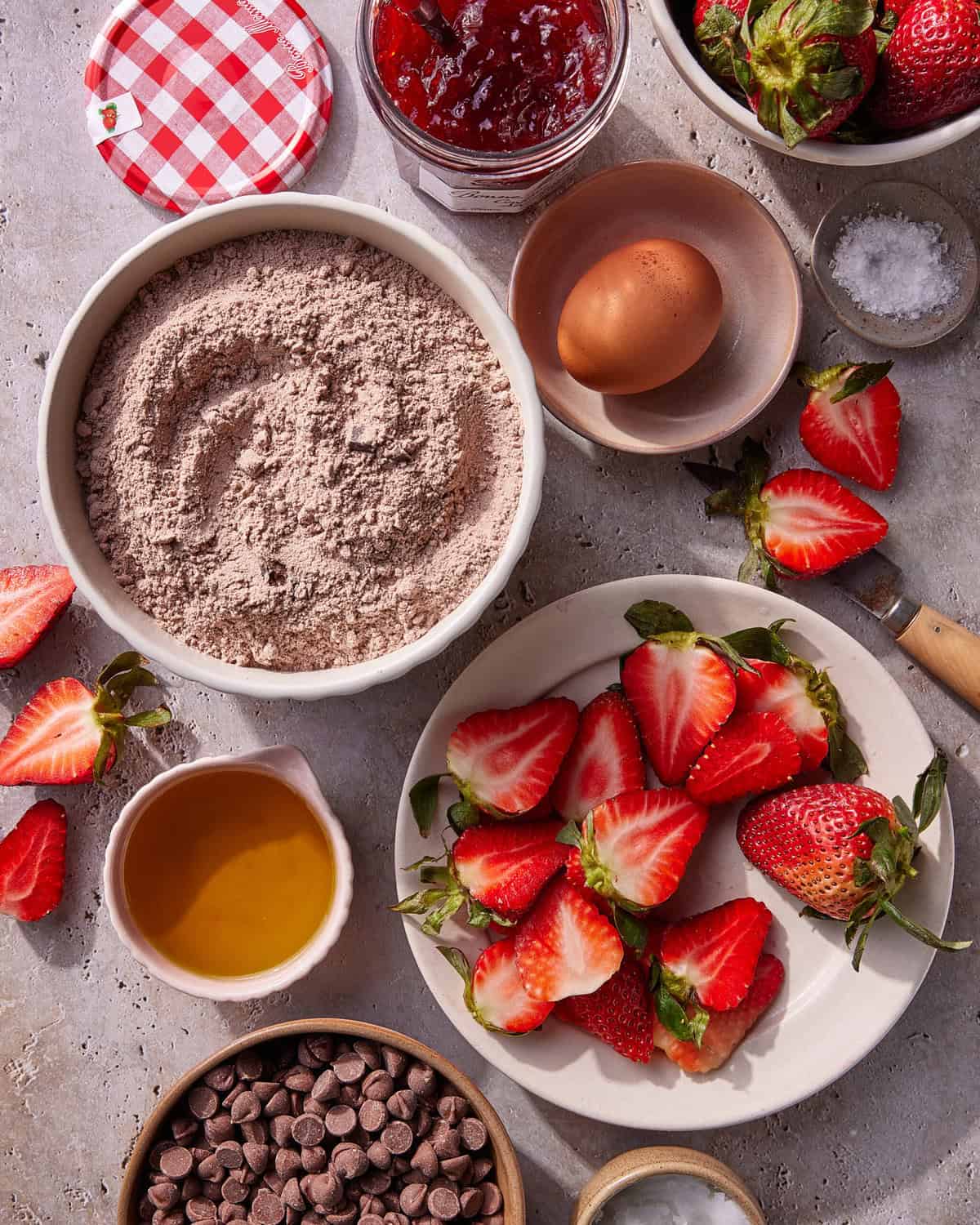 A flat lay of baking ingredients including a bowl of chocolate cake mix, a plate of halved strawberries, a cup of honey, an egg, a jar of strawberry jam, chocolate chips, salt, and whole strawberries.