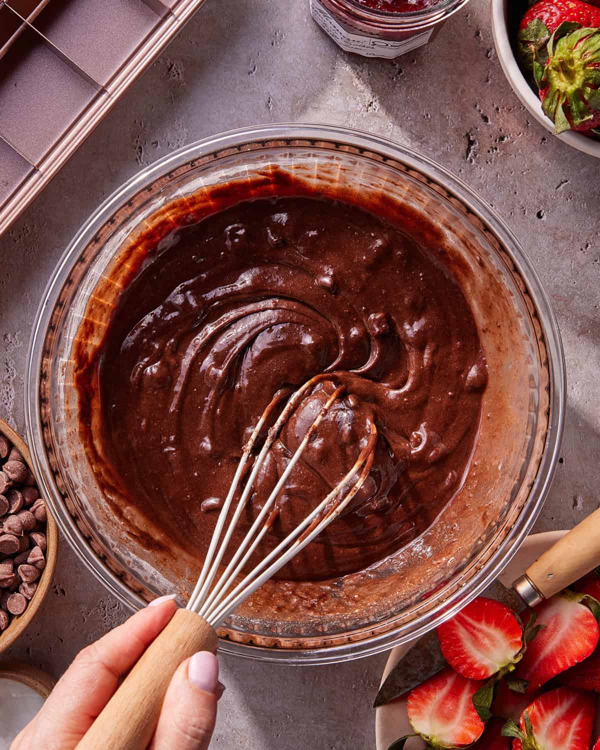 A hand holding a whisk is mixing chocolate batter in a glass bowl. Surrounding the bowl are chocolate chips, halved strawberries, a jar of jam, and a baking pan on a textured surface.