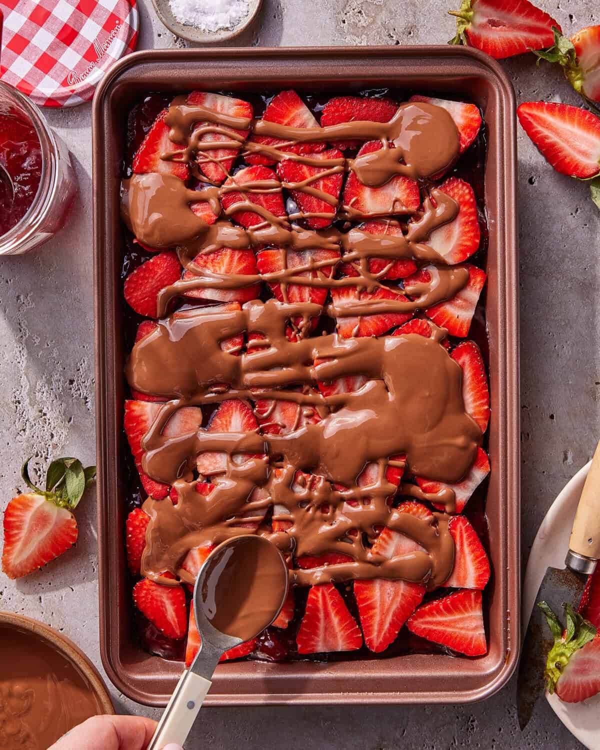 A tray of sliced strawberries topped with melted chocolate being drizzled with a spoon. A jar of jam, a knife, and strawberry stems are nearby on the table.