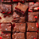 Rows of chocolate-covered strawberry brownies cut into squares, topped with flaky sea salt, and arranged on parchment paper. Some strawberry pieces and chocolate are visible at the edges of each brownie.