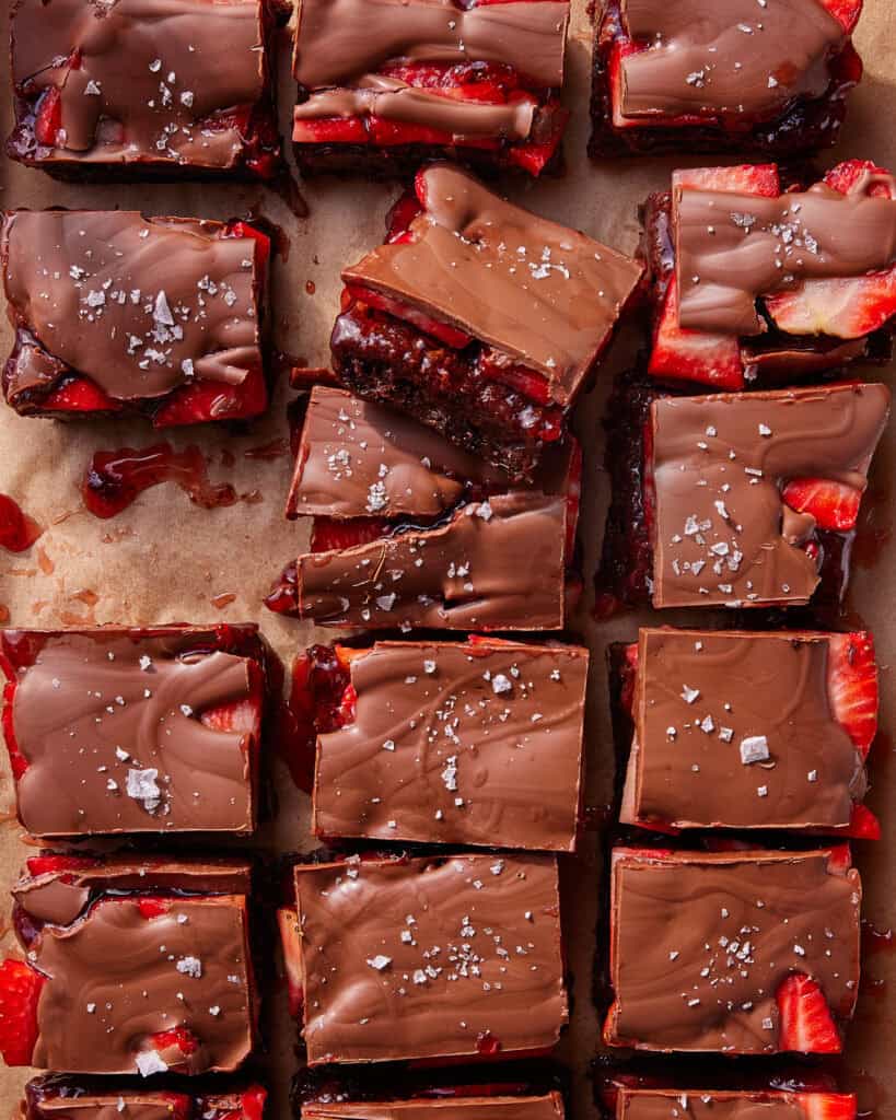Rows of chocolate-covered strawberry brownies cut into squares, topped with flaky sea salt, and arranged on parchment paper. Some strawberry pieces and chocolate are visible at the edges of each brownie.