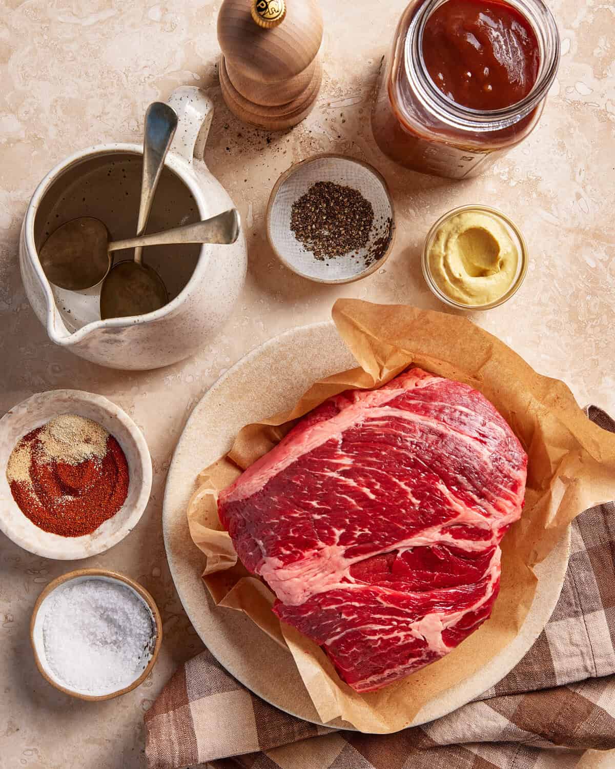 Raw marbled beef on parchment paper surrounded by bowls of spices, salt, black pepper, mustard, barbecue sauce, and a ceramic pitcher with spoons, arranged on a beige surface.