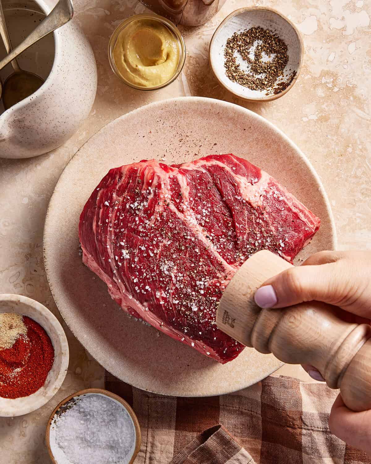 A hand seasons a raw beef roast with freshly ground pepper on a plate, surrounded by bowls of mustard, salt, ground pepper, and spices on a beige countertop.