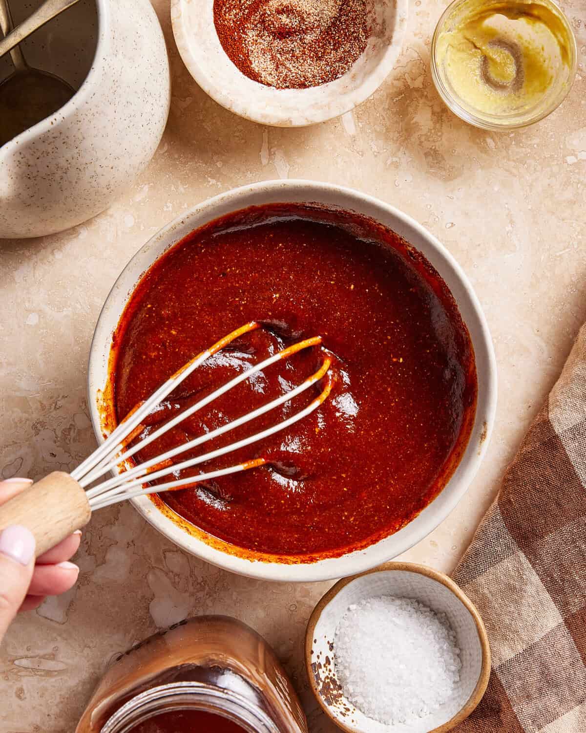 A hand holds a whisk in a bowl of red barbecue sauce, surrounded by small dishes of spices, mustard, salt, and a jar of honey on a beige countertop.