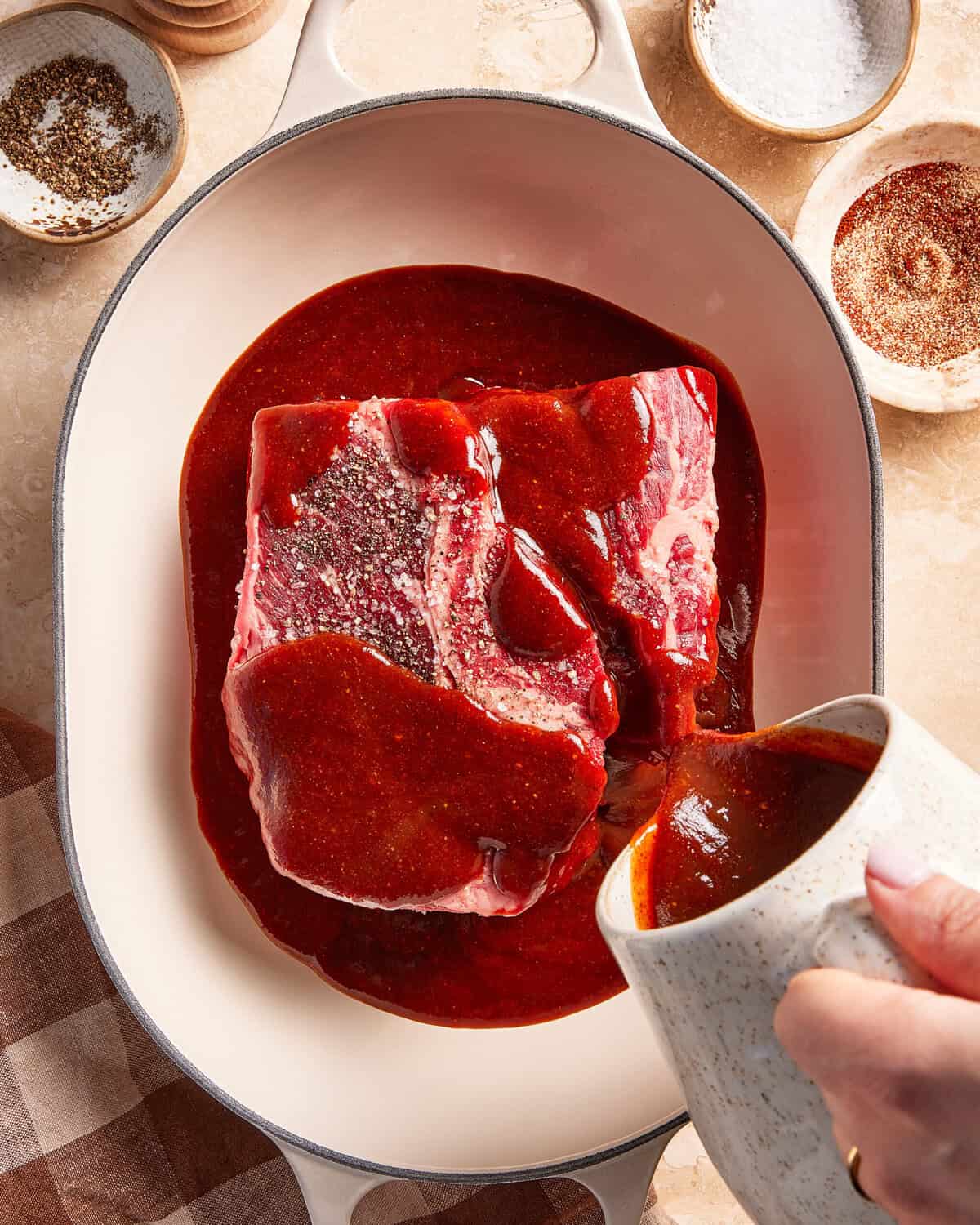 A hand pours red sauce from a white pitcher over a seasoned raw beef roast in a white oval baking dish. Small bowls of spices and salt are nearby on a tan surface.