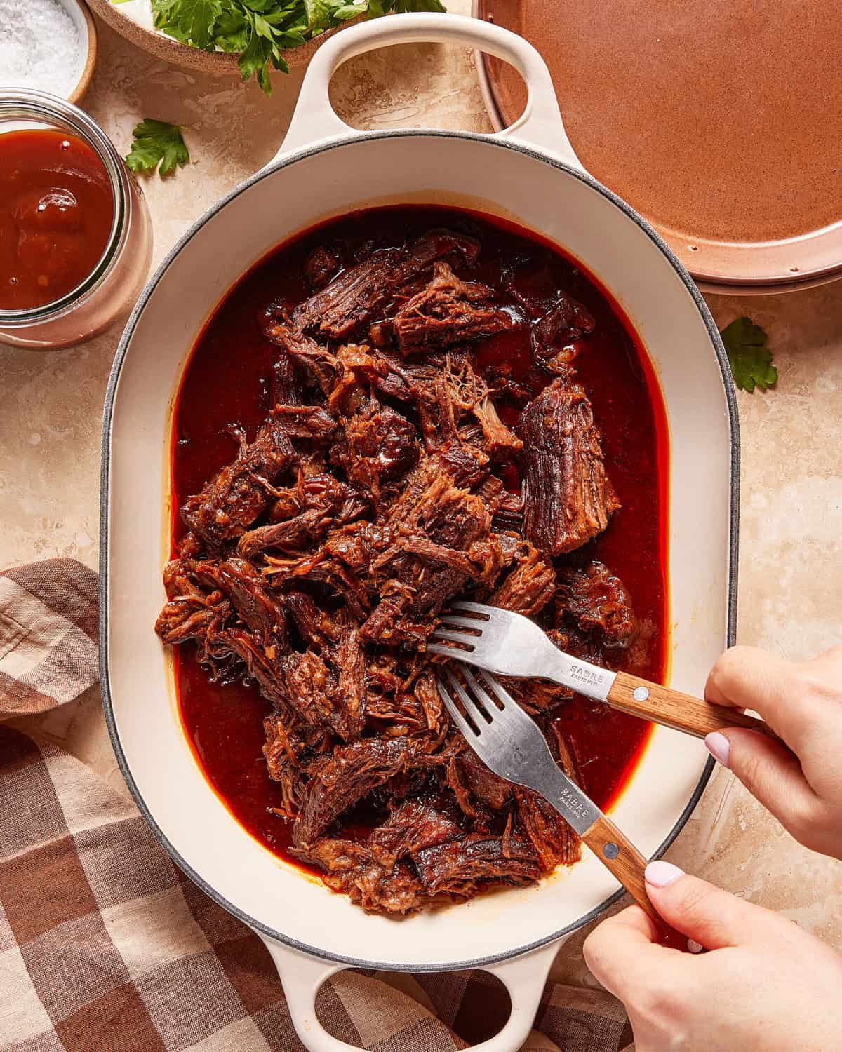 A person uses two forks to shred tender, cooked beef in a white oval dish filled with rich, red sauce. A plaid cloth, herbs, and a jar of sauce are nearby on a beige countertop.