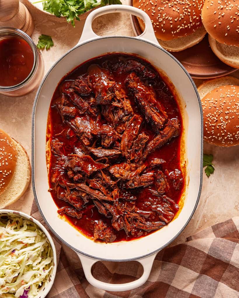 A white oval dish filled with shredded barbecue beef in a rich red sauce sits on a table, surrounded by sesame seed burger buns, coleslaw, barbecue sauce, and a checkered napkin.