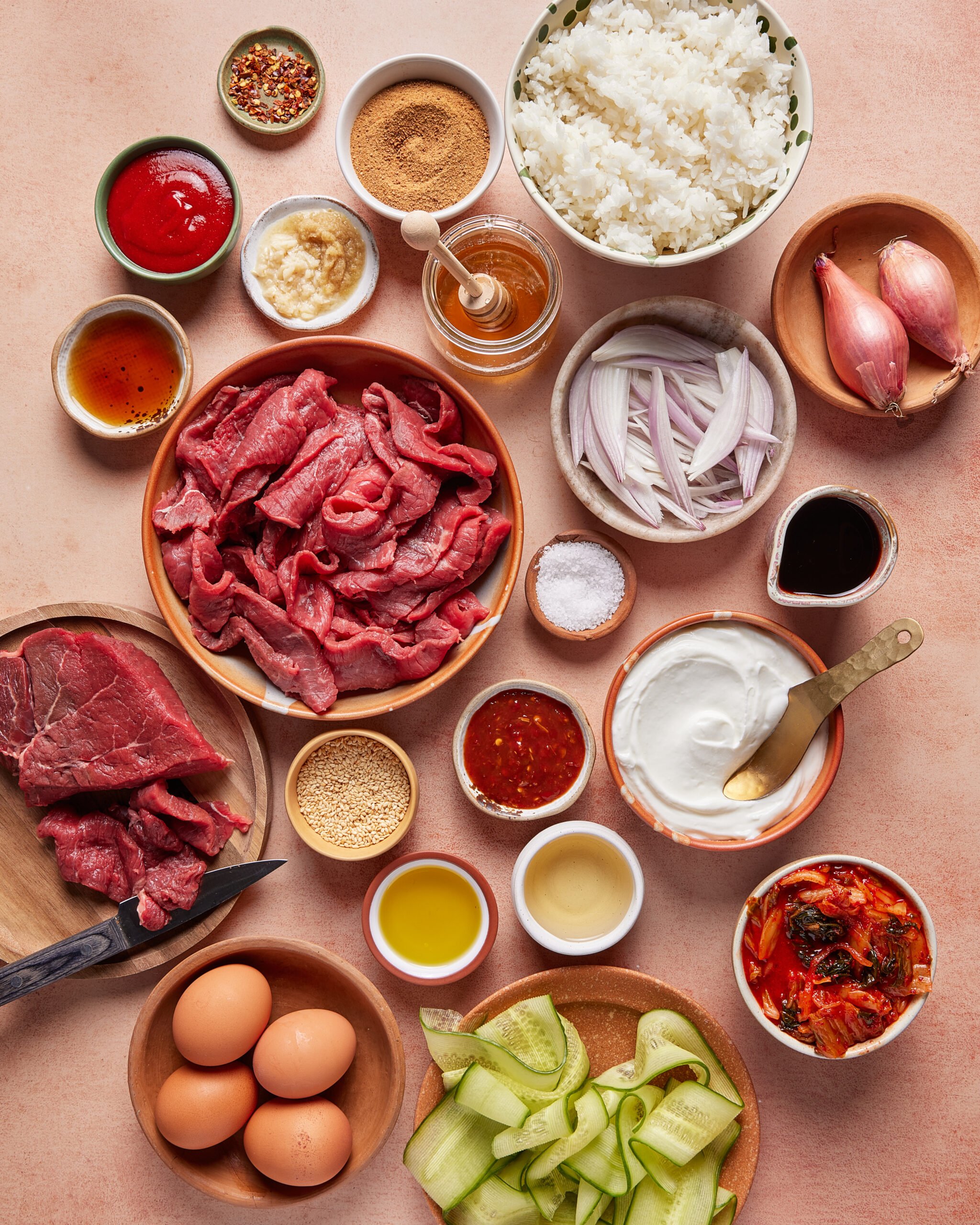 An overhead view of bowls and plates with ingredients: sliced raw beef, eggs, cooked rice, sliced onions and shallots, cucumber ribbons, kimchi, yogurt, sauces, seasonings, and a small bowl of honey, all on a peach-colored surface.