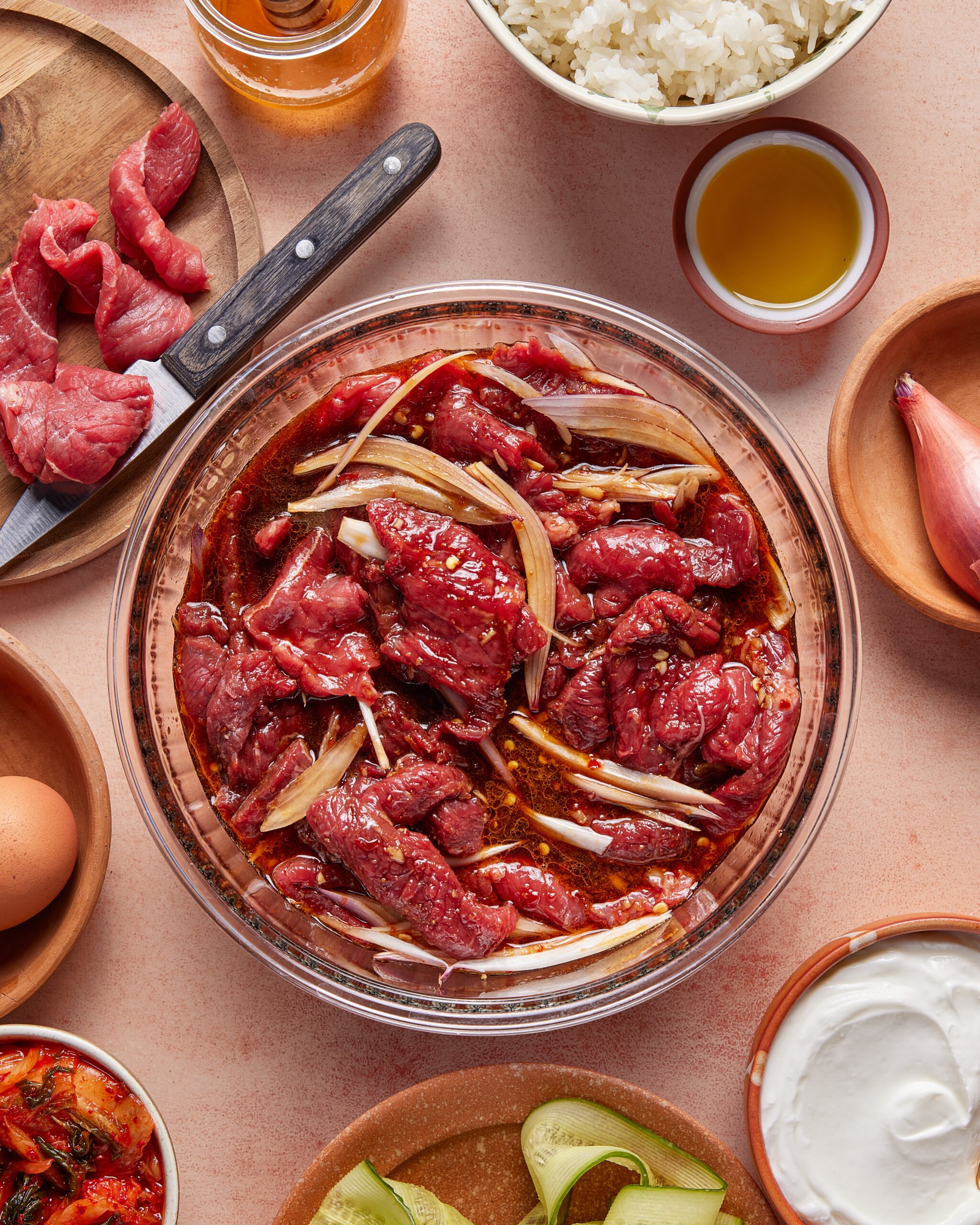 A glass bowl of marinating beef slices with onions on a table, surrounded by ingredients like olive oil, cooked rice, a raw egg, sliced cucumbers, a shallot, kimchi, and a knife with raw beef strips.