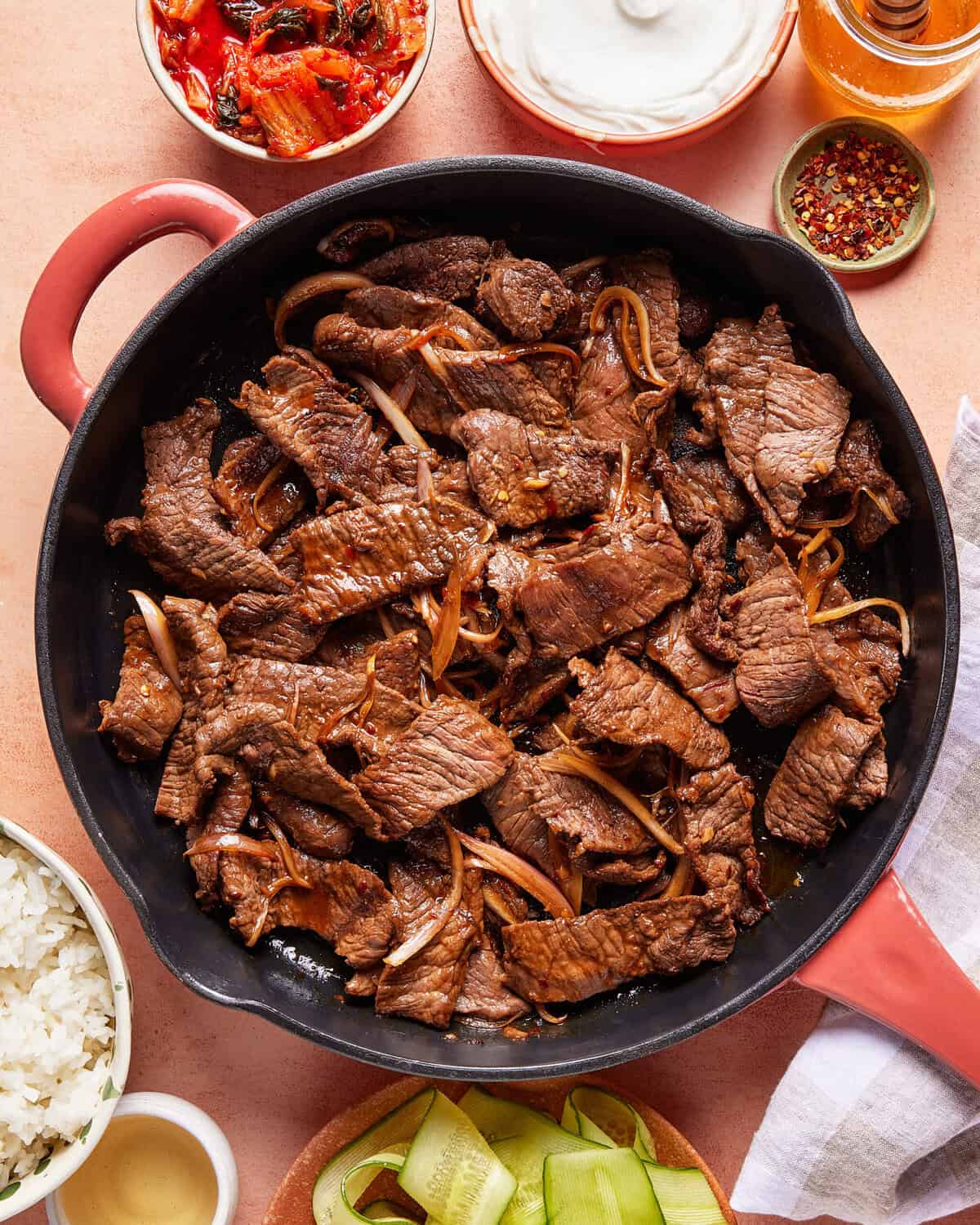 A skillet filled with cooked beef strips and onions, surrounded by bowls of rice, sliced cucumber, kimchi, yogurt, and spices on a light-colored table.