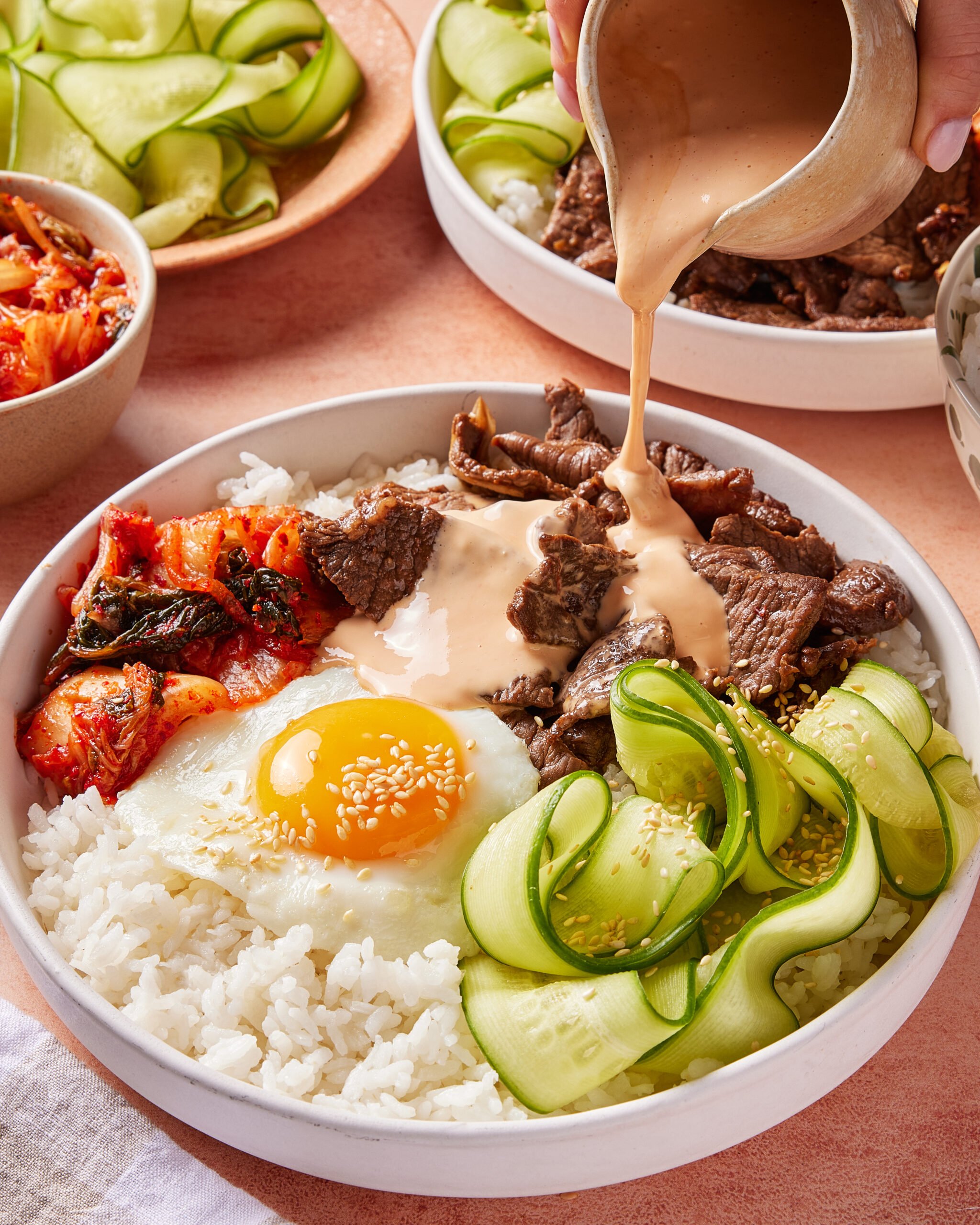A bowl filled with white rice, sliced beef, a sunny-side-up egg, kimchi, and cucumber ribbons. A hand is pouring creamy sauce over the beef. Other bowls and sides are visible in the background.