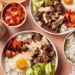 Two bowls of rice topped with grilled beef, a sunny-side-up egg, sliced cucumber, kimchi, and sauce. A small bowl of kimchi and extra rice are nearby. The meal is arranged on a peach-colored surface.