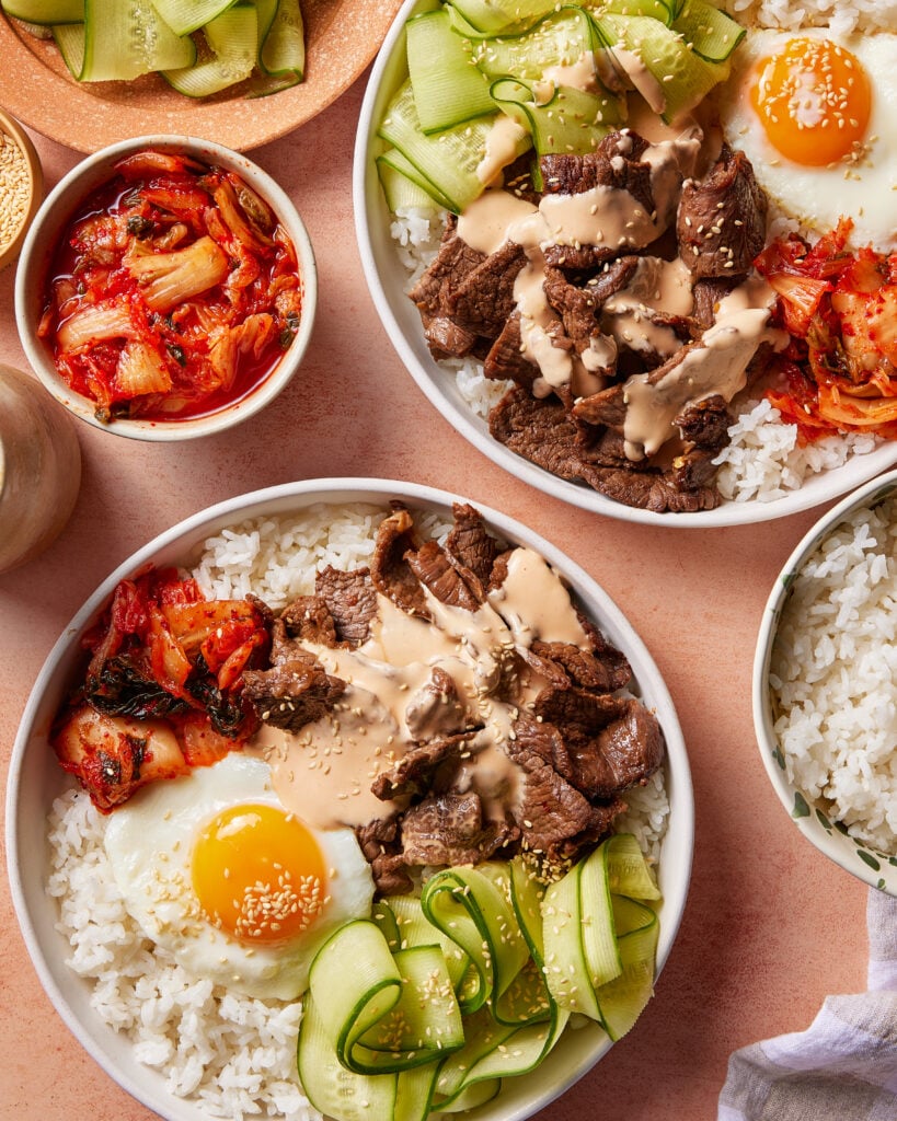 Two bowls of rice topped with grilled beef, a sunny-side-up egg, sliced cucumber, kimchi, and sauce. A small bowl of kimchi and extra rice are nearby. The meal is arranged on a peach-colored surface.