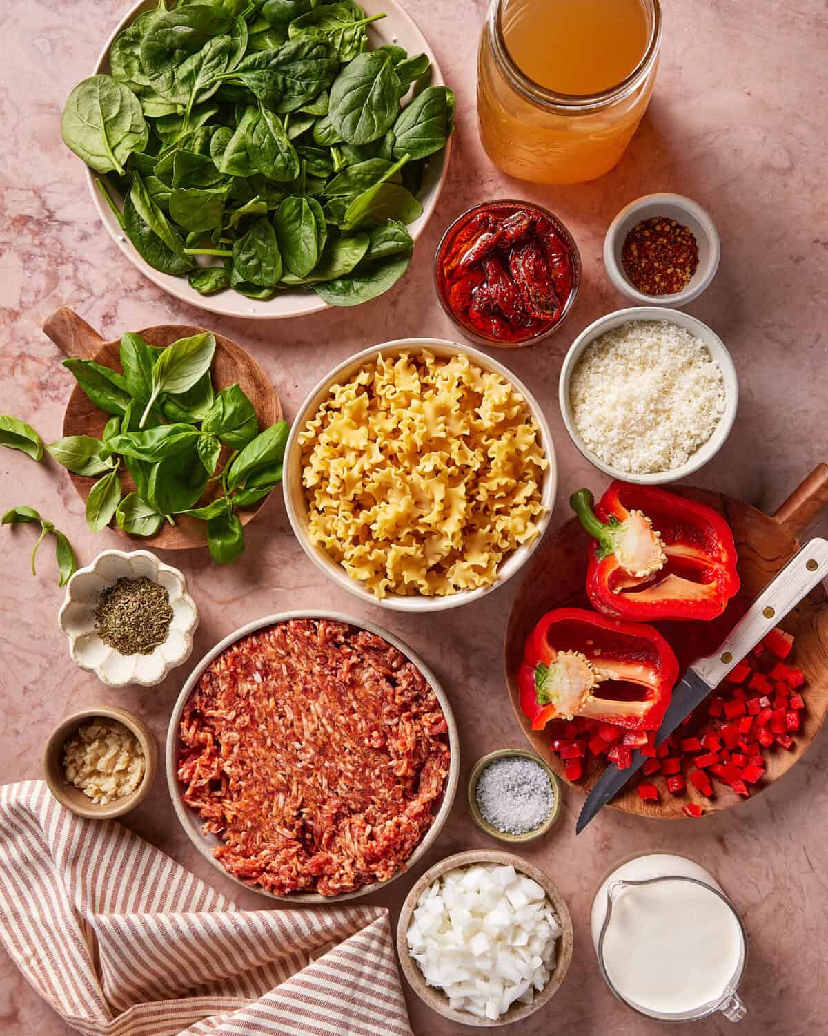 A top-down view of ingredients for a pasta dish, including ground meat, uncooked pasta, spinach, chopped red bell pepper, onion, garlic, basil, sun-dried tomatoes, grated cheese, cream, broth, and seasonings on a counter.