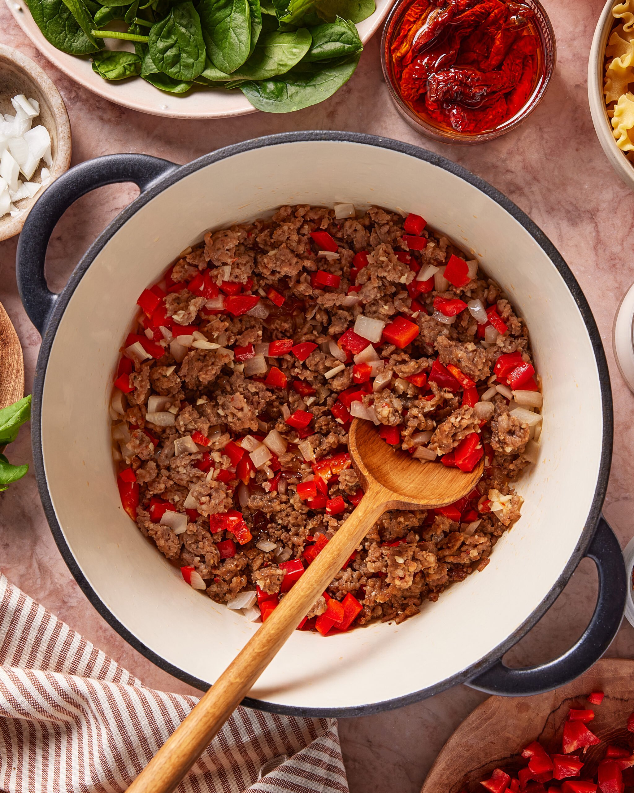 A pot of cooked ground meat with chopped red bell pepper and onion, stirred with a wooden spoon. Surrounding the pot are bowls of fresh spinach, sun-dried tomatoes, raw pasta, and more chopped onions.