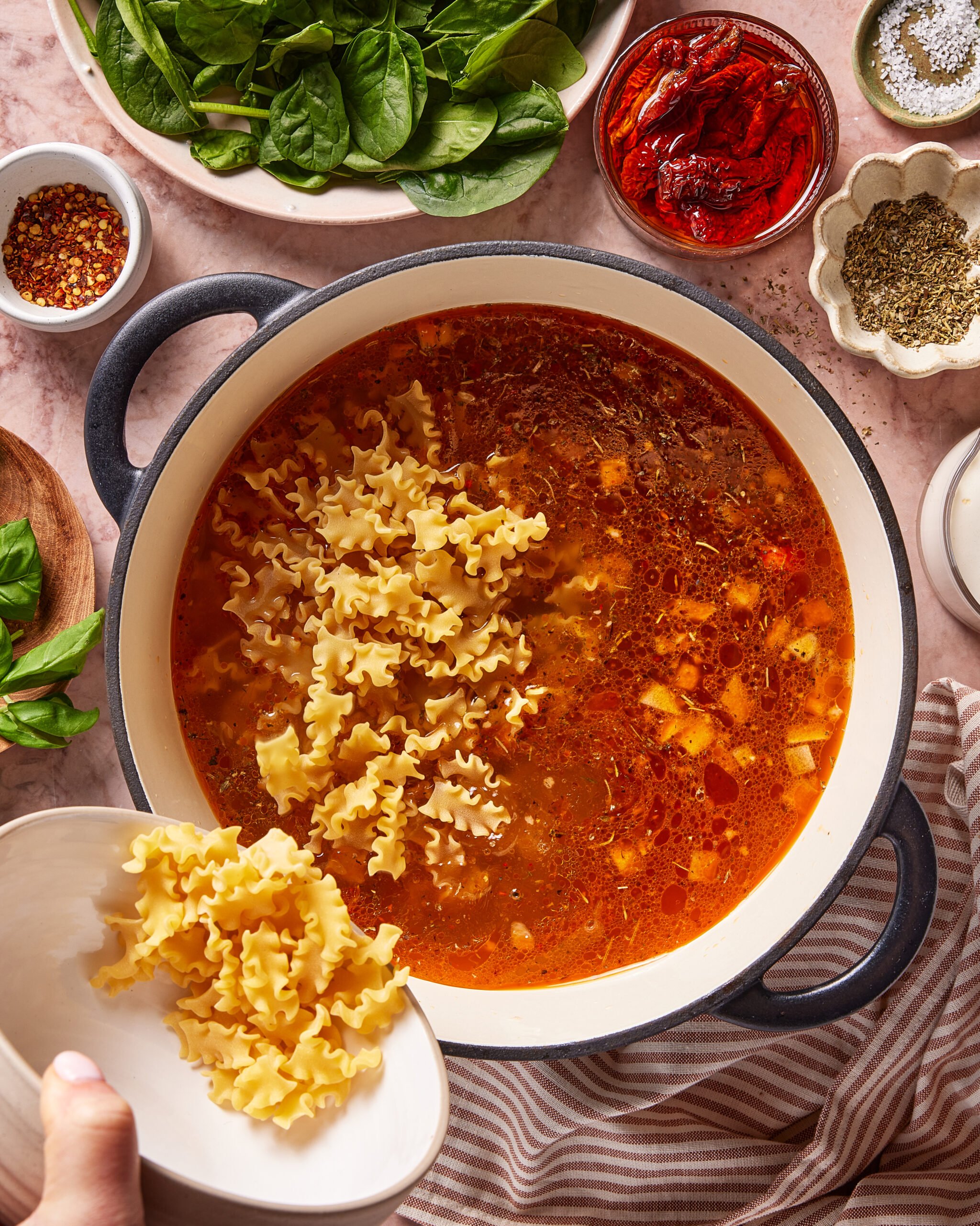 A hand pours uncooked mafaldine pasta into a pot of tomato-based soup on a countertop, surrounded by bowls of spinach, herbs, red pepper flakes, sun-dried tomatoes, and seasonings.