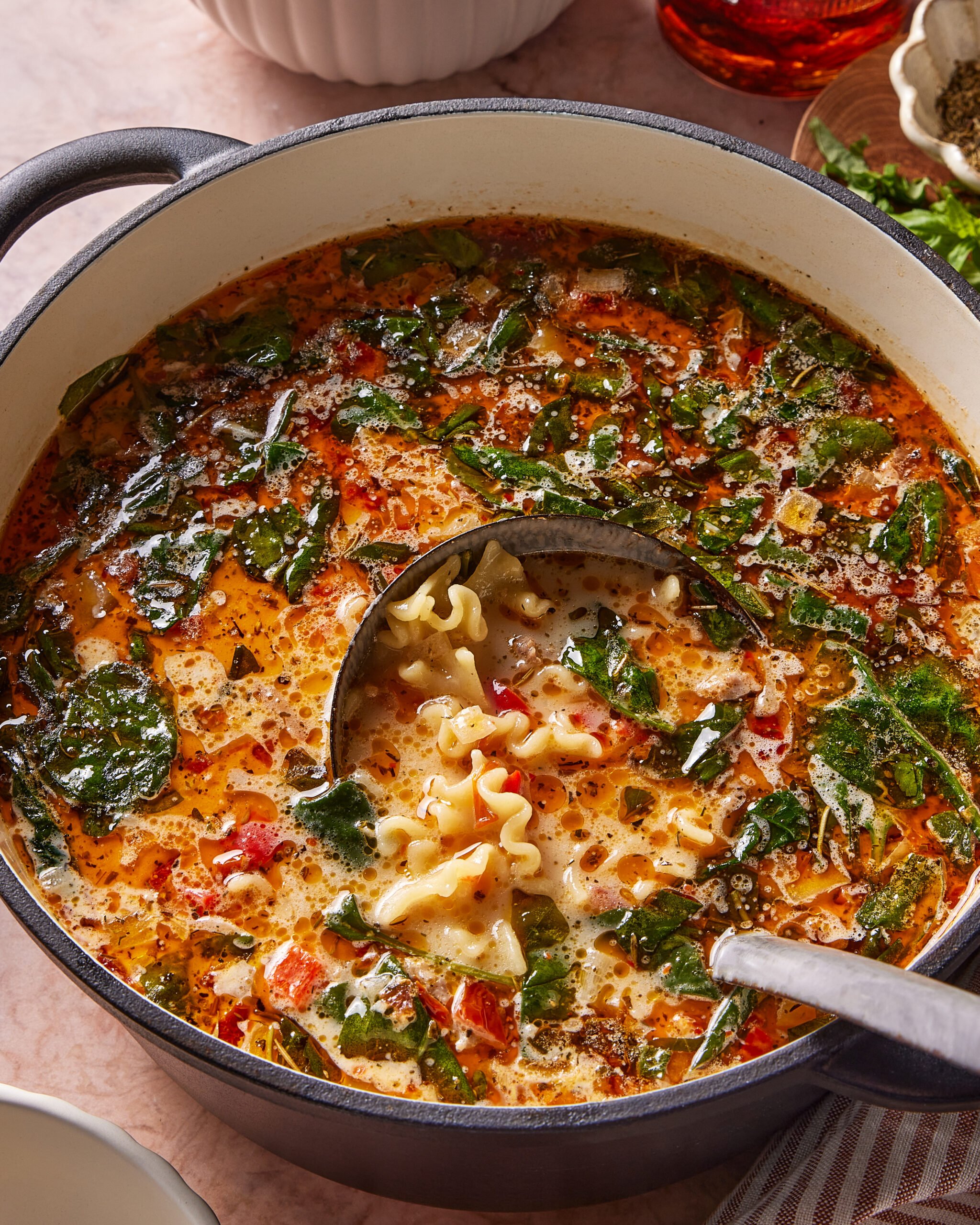 A large pot of soup filled with curly pasta, spinach, diced tomatoes, and herbs. A ladle is scooping up a serving, showing the ingredients in a rich, savory broth.