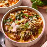 A bowl of lasagna soup with curly noodles, spinach, tomatoes, and ground meat, topped with grated cheese and fresh basil, sits on a pink surface beside a spoon and a plate of herbs.