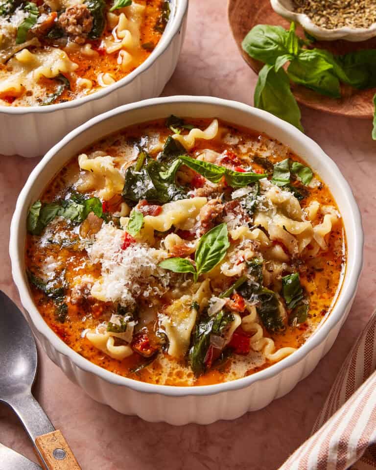 A bowl of lasagna soup with curly noodles, spinach, tomatoes, and ground meat, topped with grated cheese and fresh basil, sits on a pink surface beside a spoon and a plate of herbs.
