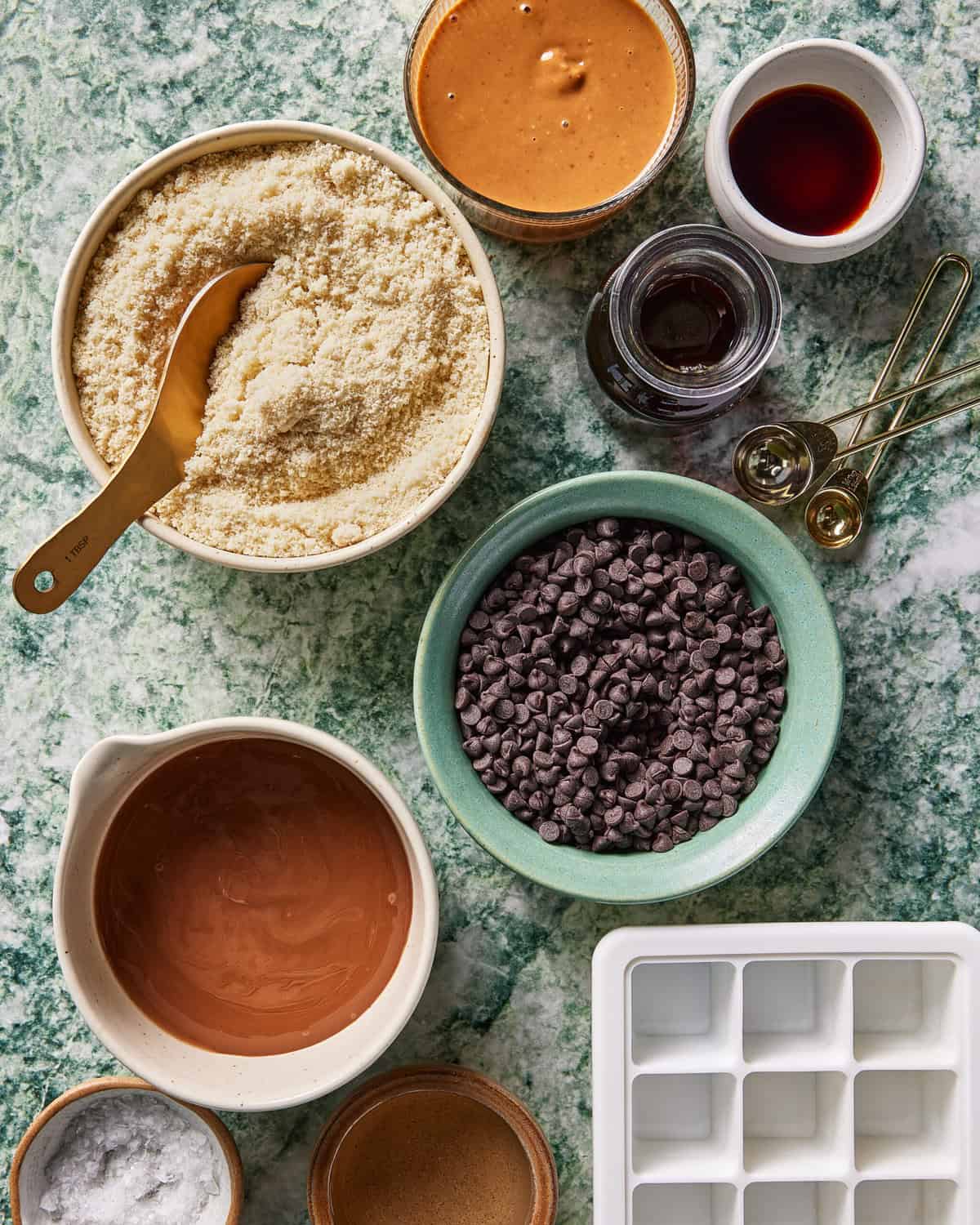 Bowls with almond flour, chocolate chips, melted chocolate, peanut butter, vanilla extract, and measuring spoons are arranged on a green marble countertop next to an empty white ice cube tray.