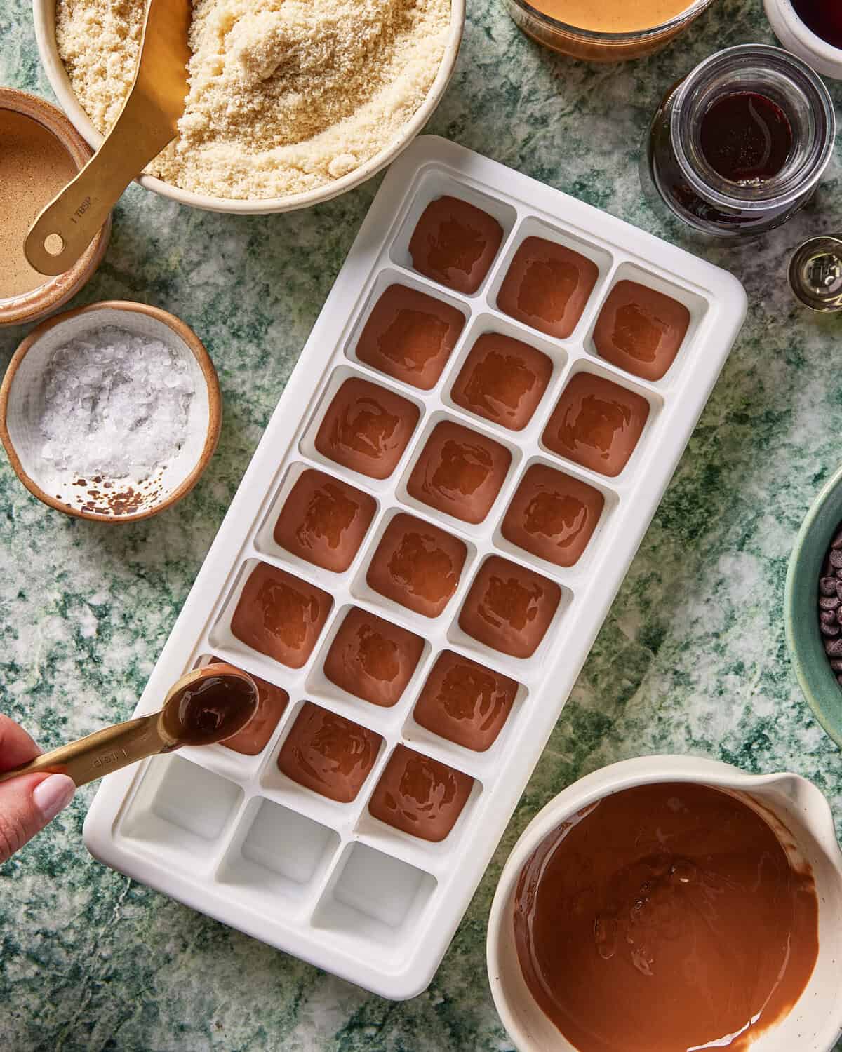 A hand fills an ice cube tray with melted chocolate, surrounded by bowls of almond flour, salt, and melted chocolate on a green marbled countertop.