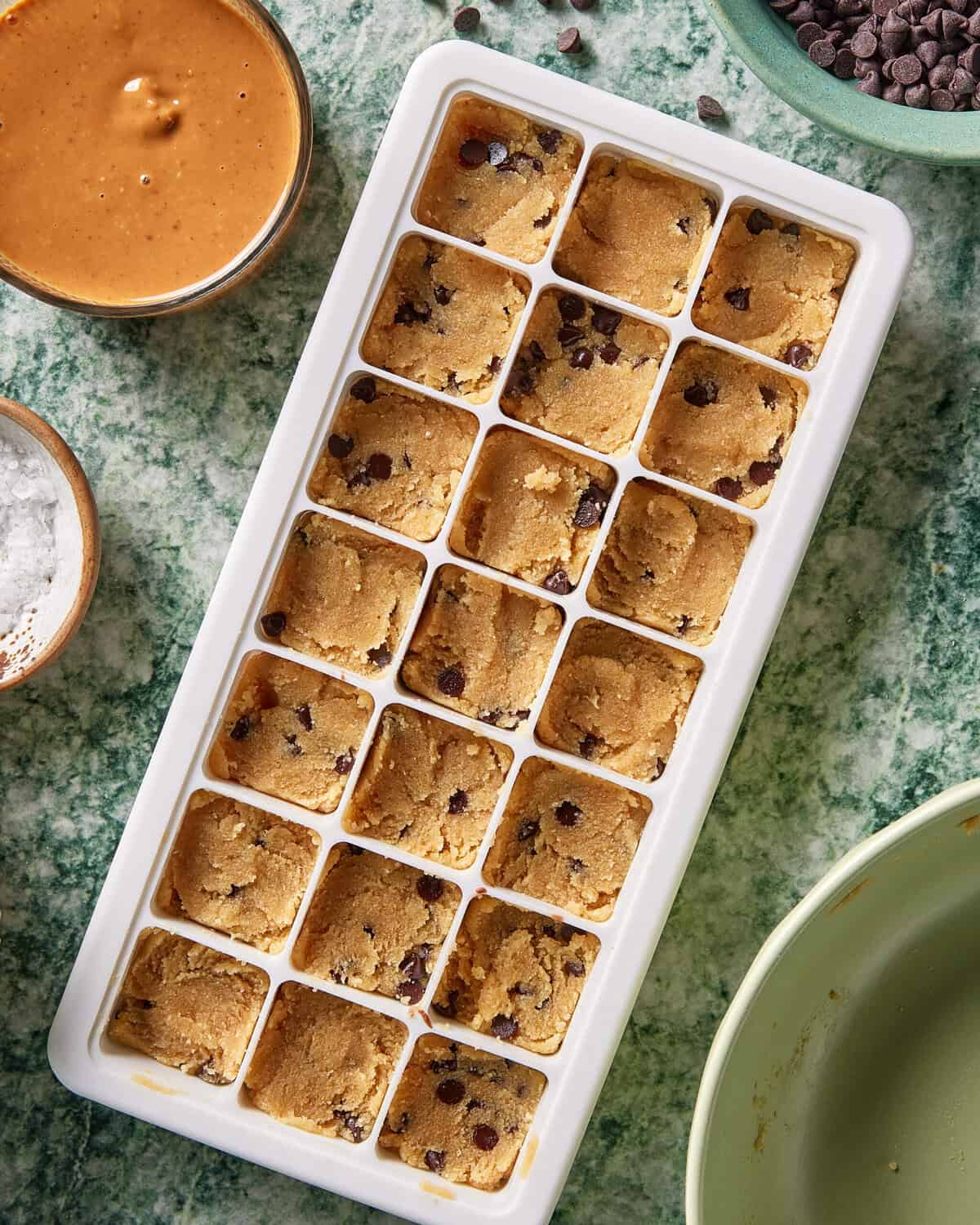 A white ice cube tray filled with balls of chocolate chip cookie dough sits on a green countertop, surrounded by bowls of peanut butter, sea salt, and chocolate chips.