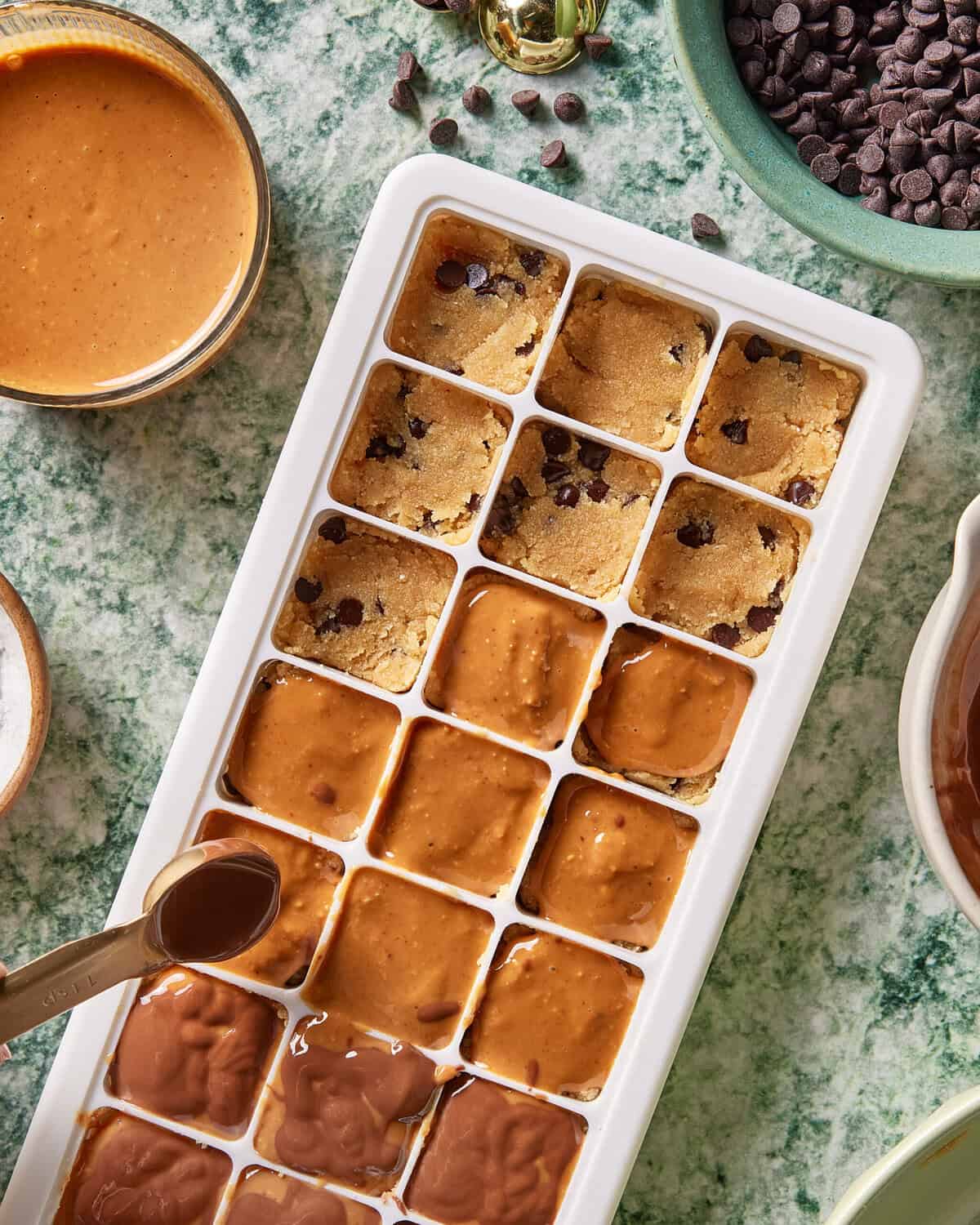 An ice cube tray filled with sections of chocolate chip cookie dough and a creamy brown mixture, with bowls of chocolate chips, peanut butter, and a spoon nearby on a green marble surface.