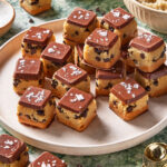 A plate filled with square, layered cookie dough bars topped with chocolate and a sprinkle of sea salt, surrounded by bowls of chocolate chips and ingredients on a green marble surface.