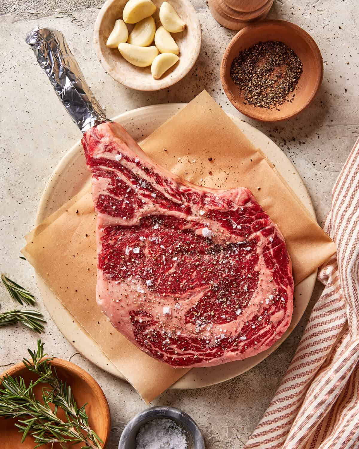 Raw, seasoned tomahawk steak on parchment paper, surrounded by bowls of garlic cloves, ground black pepper, rosemary sprigs, and salt, with a striped cloth on a stone surface.